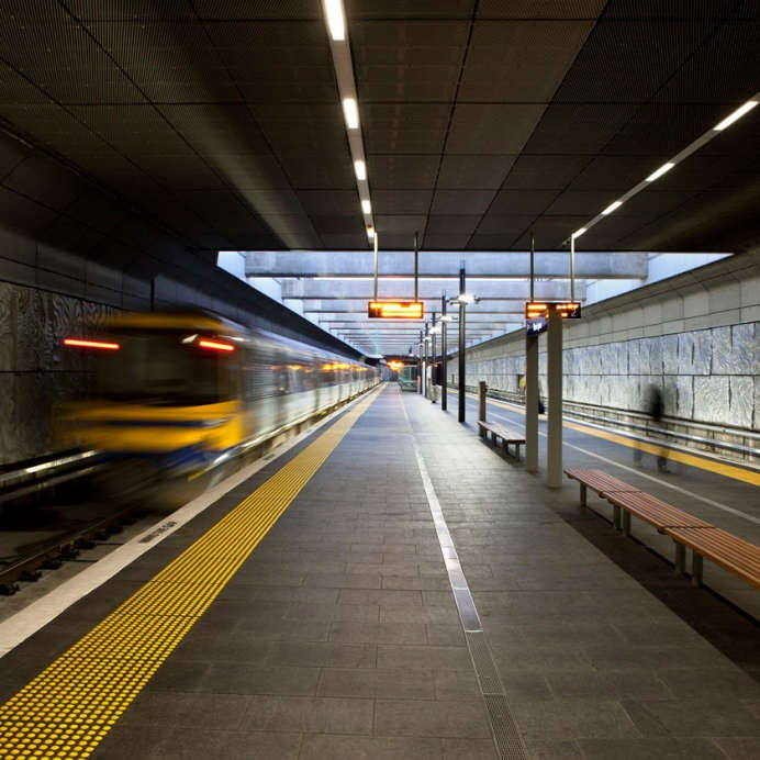 Blurry train disappears away from viewer in underground train station platform. TacPro yellow polyurethane tactile indicators installed along platform edge on both sides