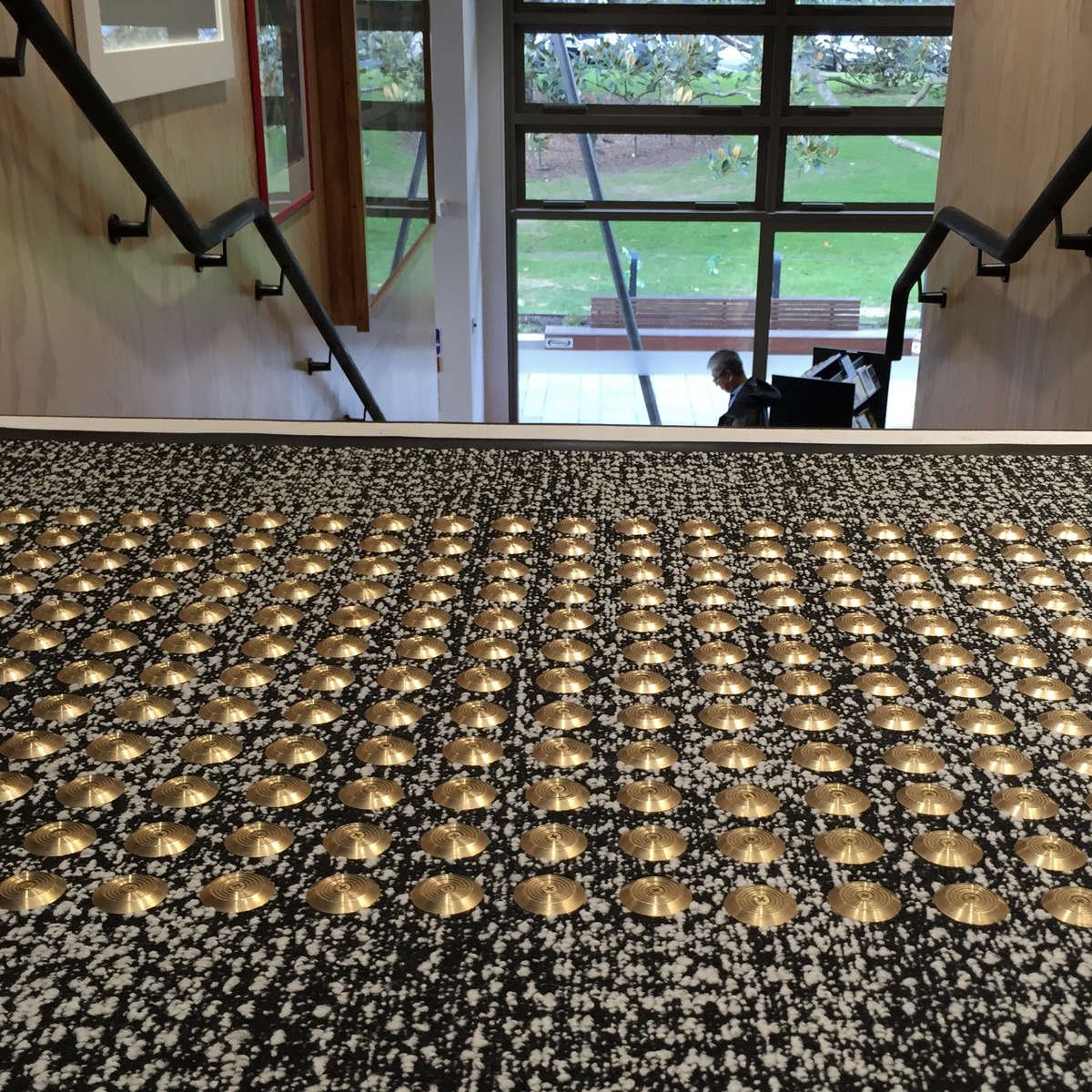 TacPro brass tactile indicators installed on black carpet with white speckles at top of stairs