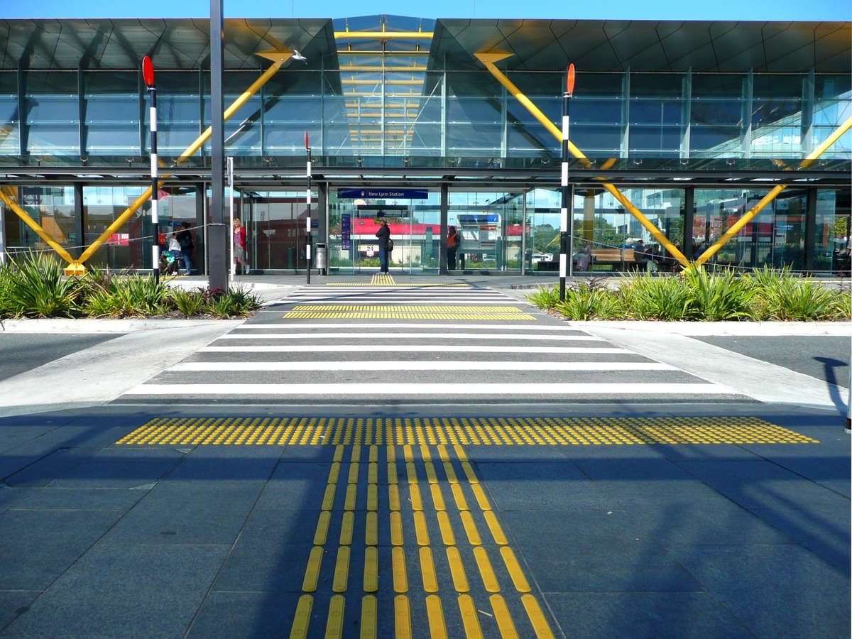 TacPro yellow polyurethane warning and directional tactile indicators on bluestone tile at large pedestrian crossing (with mid-block), leading to New Lynn Train Station main building