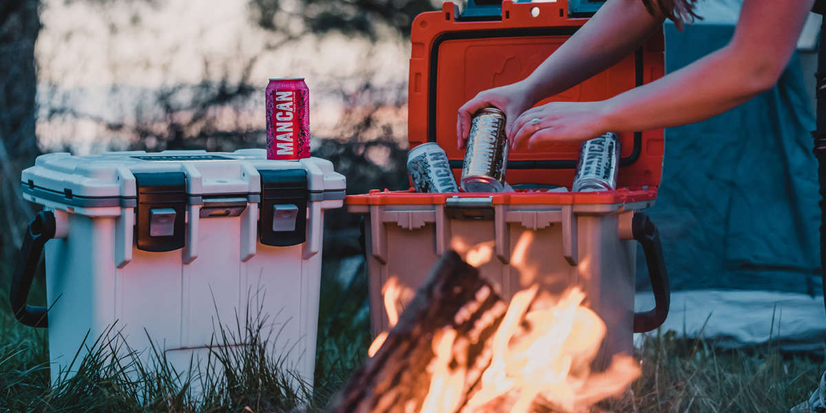 Two Pelican Elite Coolers side by side near a campfire; one is closed and the other is open. A person is pulling a canned beverage out of the ice in the open cooler.