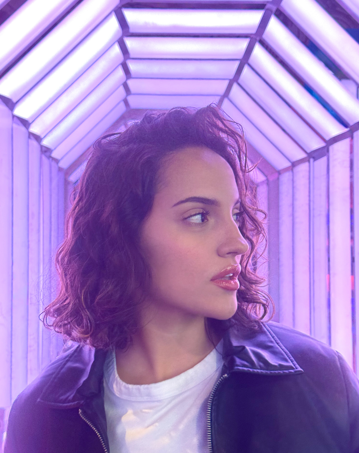 Women in a white t shirt and black leather jacket in front of purple lit hallway.