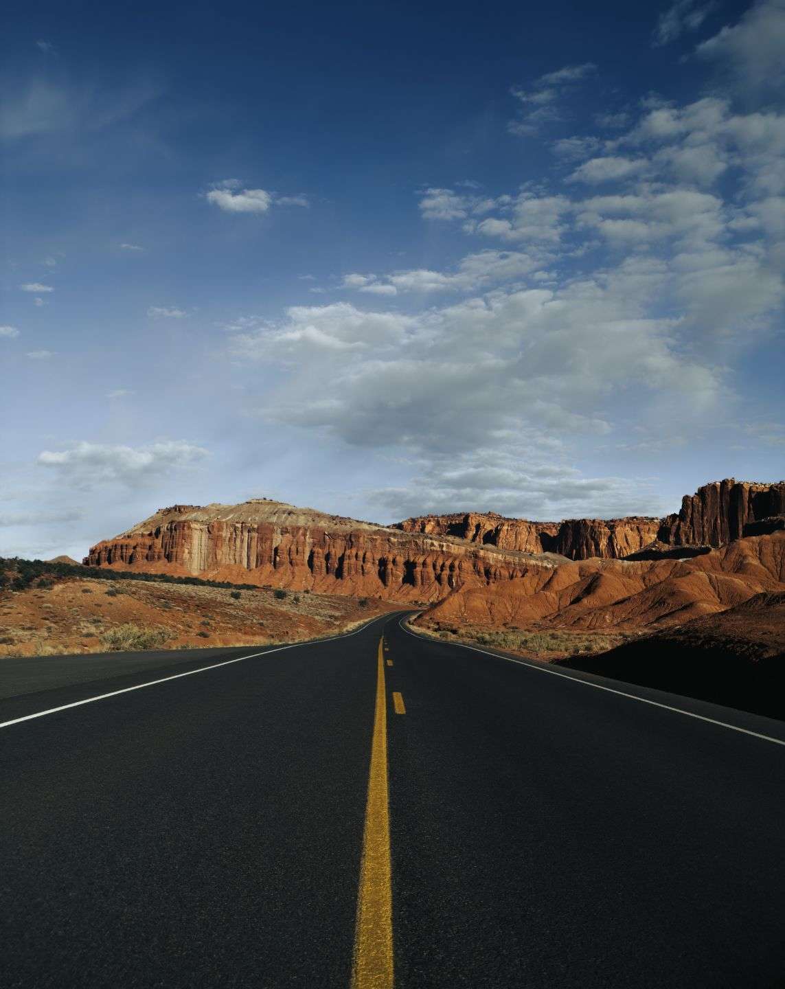 Desert highway leading toward red rock cliffs under a blue sky with scattered clouds, captured on iPhone using SANDMARC gear — showcasing strong leading lines and dynamic perspective in mobile photography.