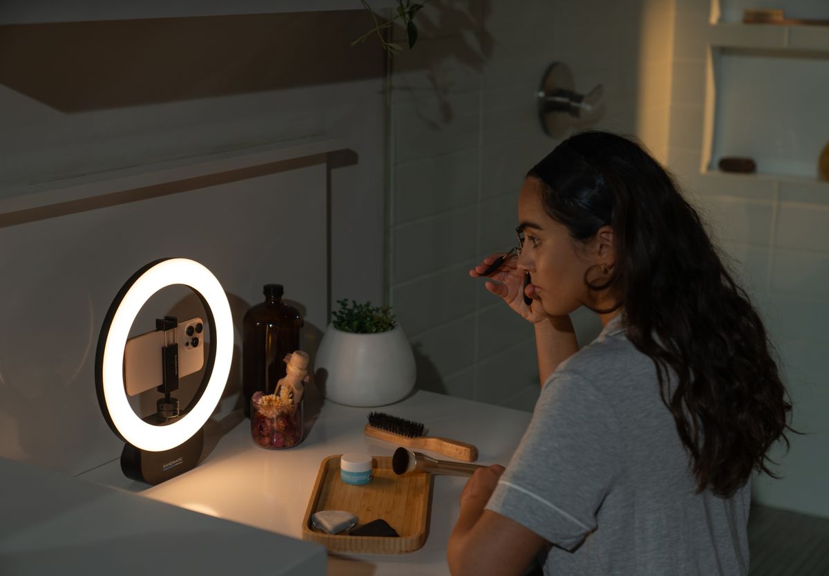 Woman putting on mascara at a vanity looking towards an iPhone attached to a SANDMARC Ring Light.