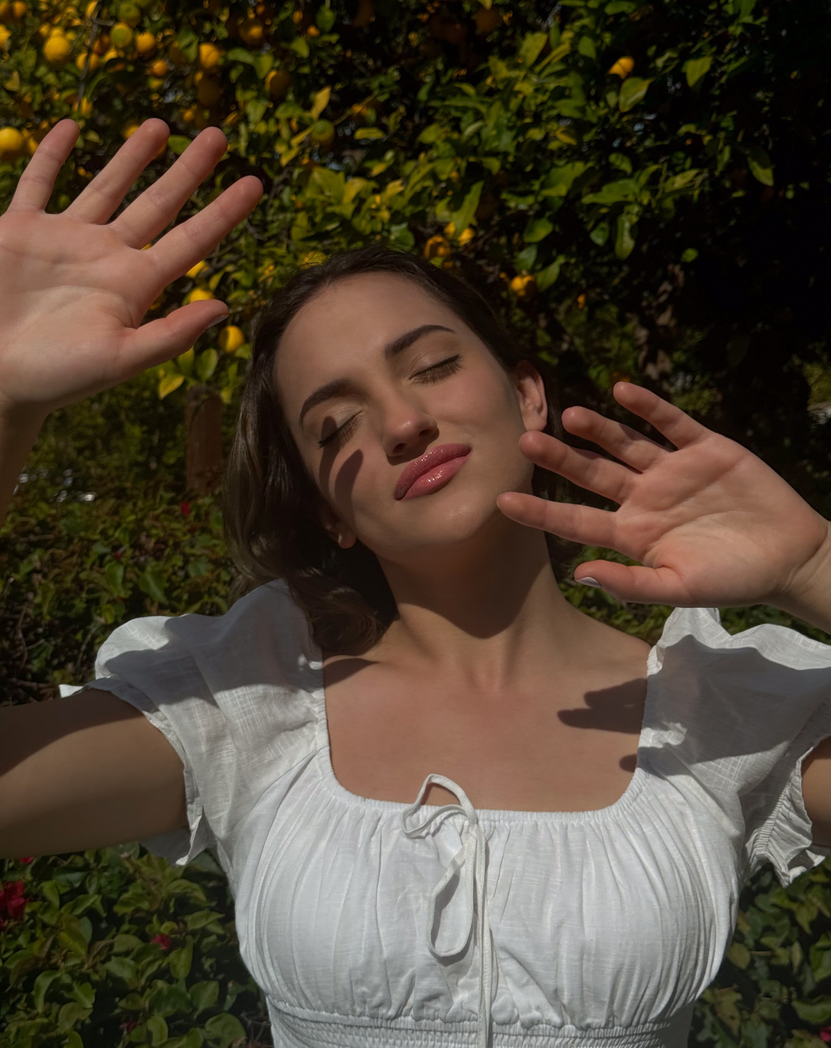 Women in a white top with hands slightly over face blocking sunlight, with a lemon tree in background.