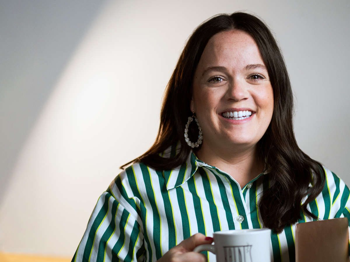 A woman smiles broadly as she holds a cup of coffee and a clipboard