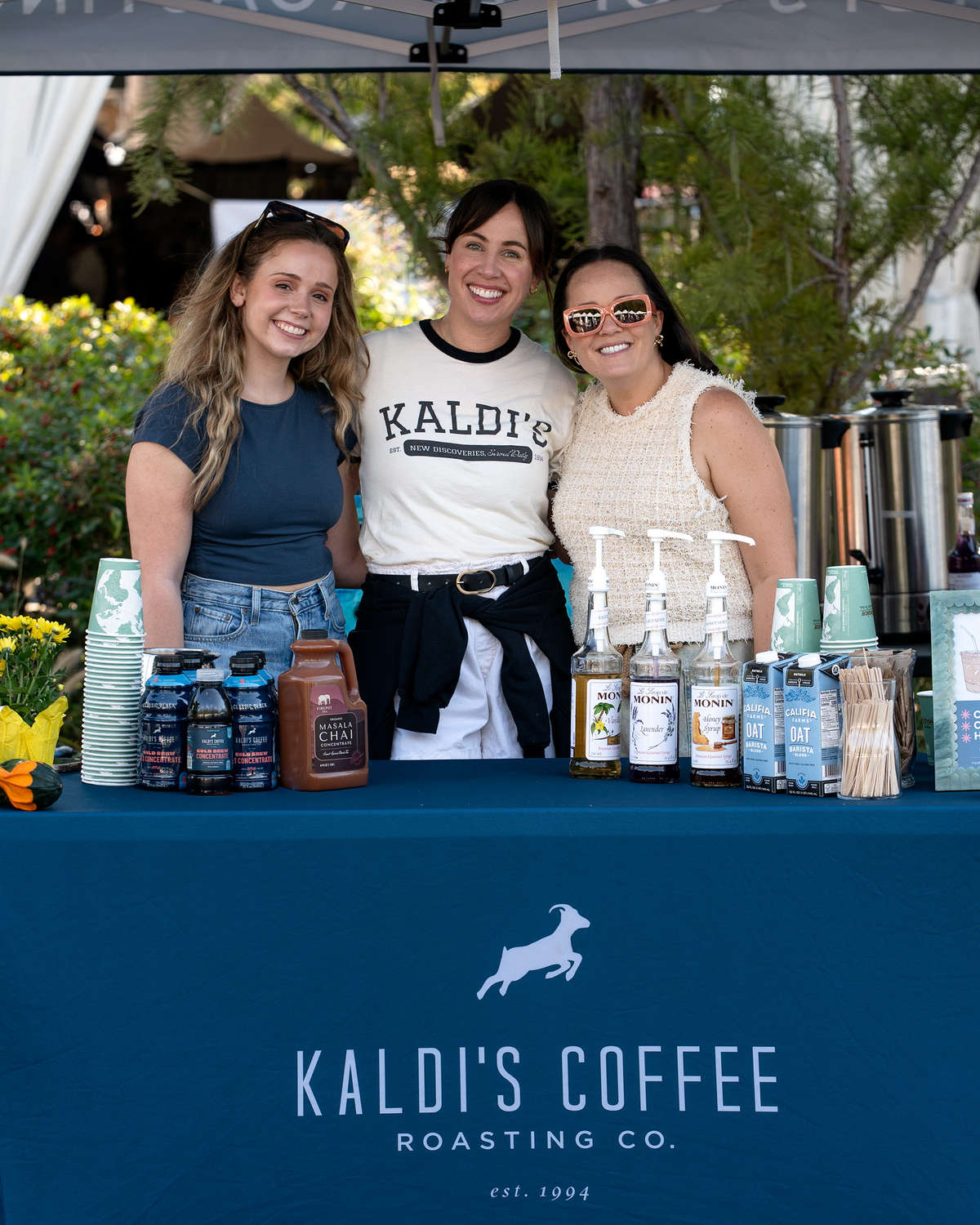 Three brunette woman stand behind a Kaldi's branded table, arm in arm, smiling broadly