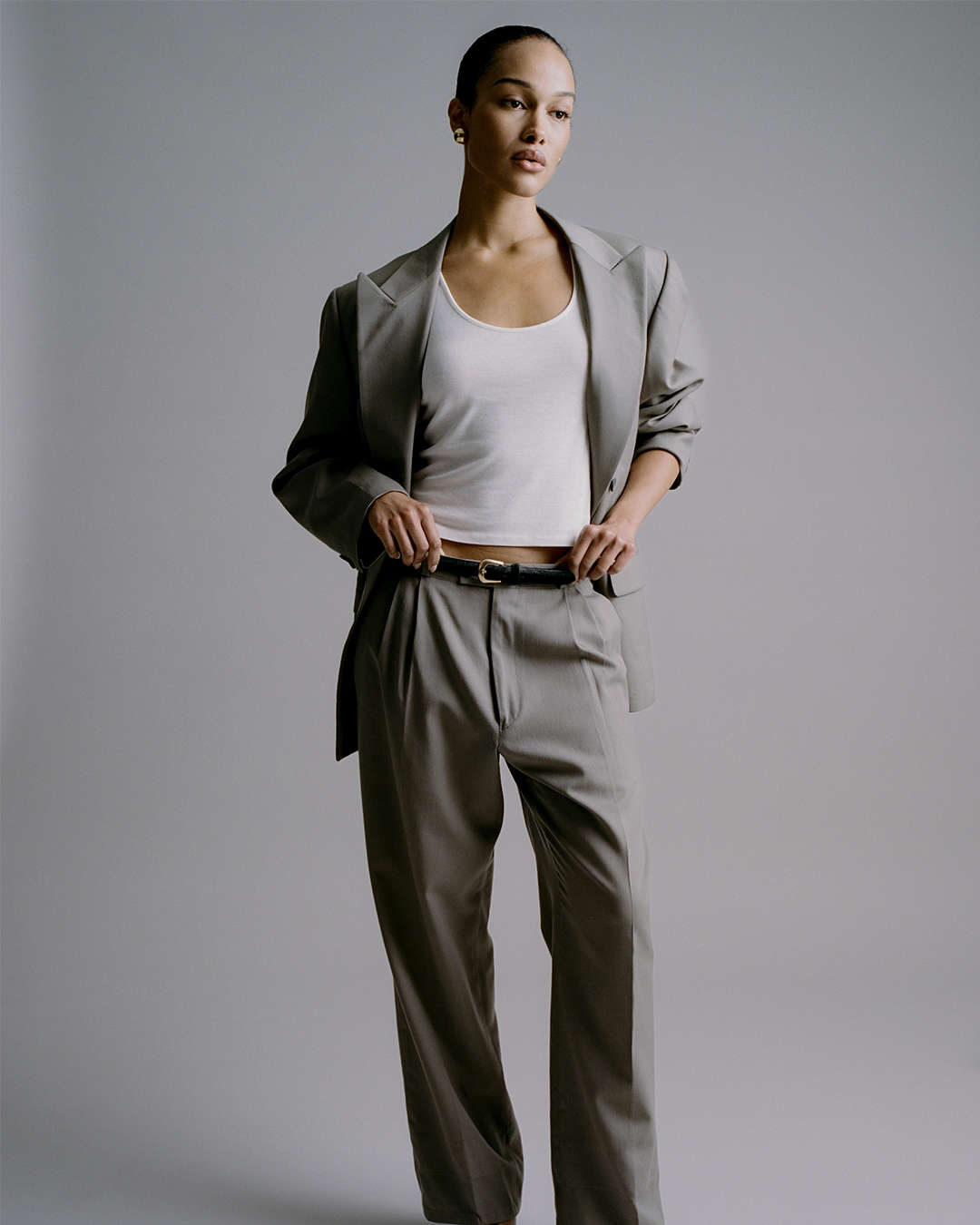 model against a gray backdrop wearing a grey suit white top and skinny black calf-hair belt