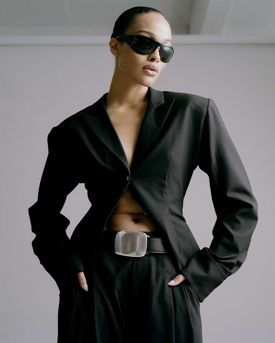 model wearing a black suit and black sunglasses with a black leather belt that has an oversized silver plaque buckle