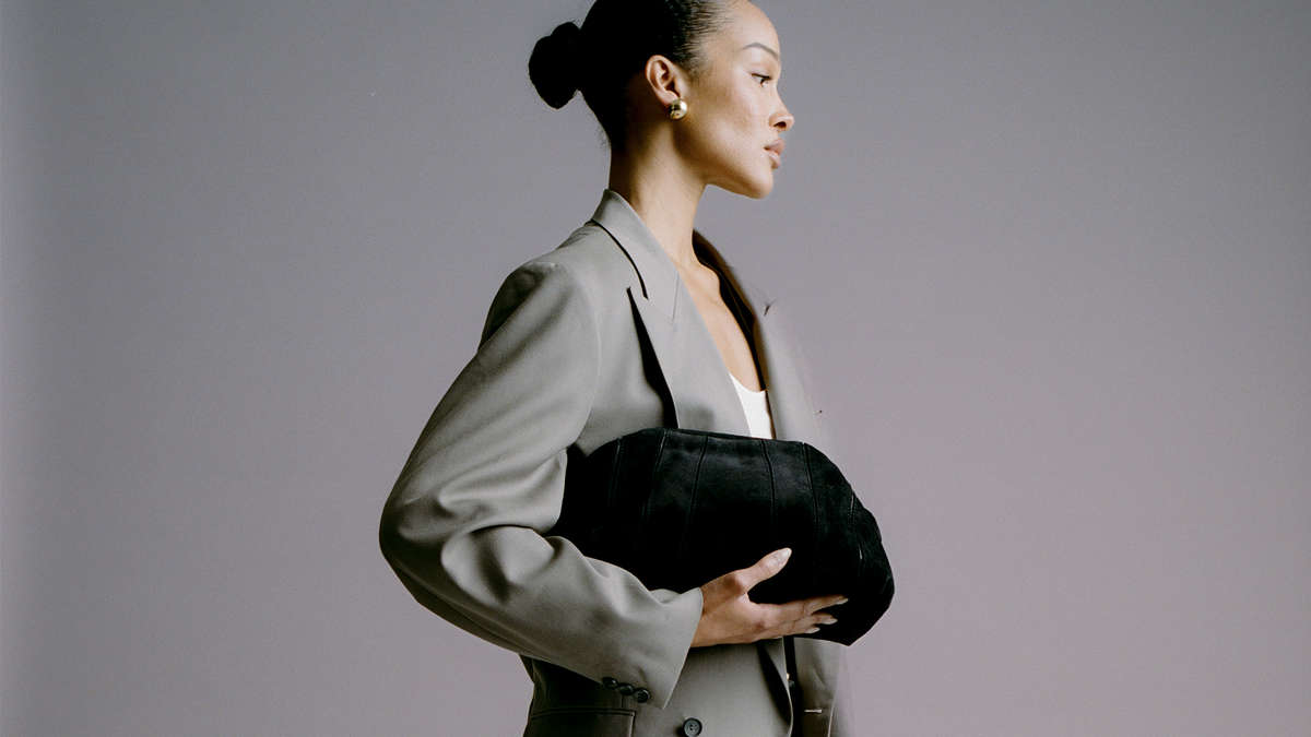model against a grey backdrop wearing a light grey suit and holding a black suede clutch