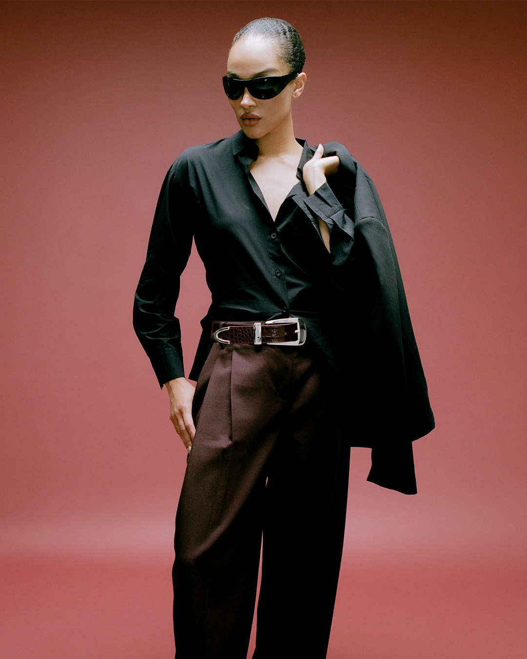 model agaisnt a burgundy backdrop wearing a black top with burgundy trousers and a burgundy croc-print belt with oversized silver hardware