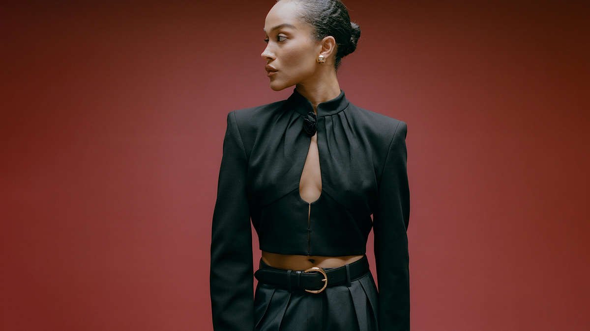 model against a burgundy backdrop with a black top, black leather belt with a gold buckle, and dark trousers 